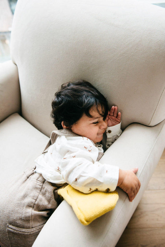 A toddler with curly hair is lying on a white couch, playfully leaning against the cushion. Clad in a white shirt and brown overalls, they snuggle next to a small yellow pillow. The room's natural light creates the perfect setting for charming commercial photography.