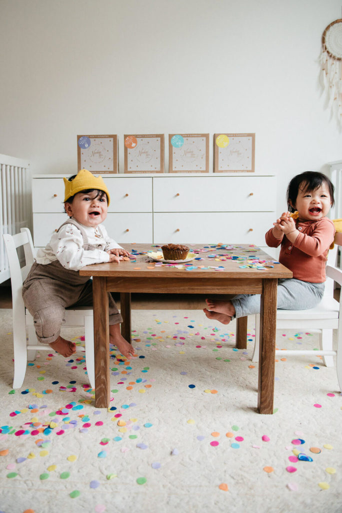 Two toddlers sit at a small table, surrounded by colorful confetti on the floor, perfectly capturing the charm of brand photography. One wears a yellow hat and beige overalls; the other dons a rust-colored top. A cupcake awaits on the table, framed by art-filled walls.