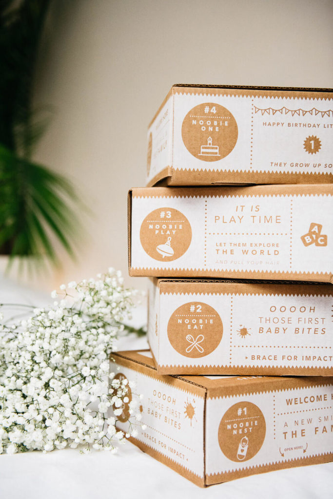 Stack of four playful cardboard boxes featuring baby toys and gifts, captured in an elegant brand photography style. A small bunch of white flowers beside the boxes, with a plant adding a touch of greenery, completes this charming commercial display.