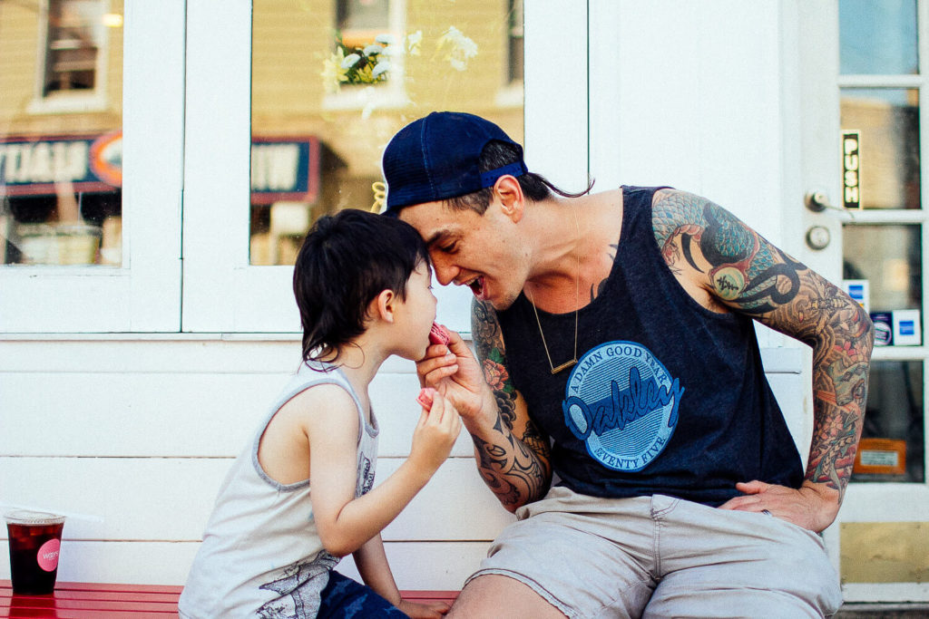A tattooed man in a cap playfully leans forward to touch noses with a young child, capturing a heartwarming moment perfect for brand photography. They're both seated outside a building, with the man holding a small drink and a larger one on the table beside them.