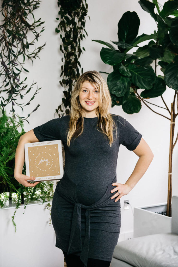 A woman in a dark gray dress stands smiling indoors, holding a brown box with Natalie written on it. Capturing the essence of brand photography, she is surrounded by lush green plants, including a tall fiddle leaf fig and cascading ferns.
