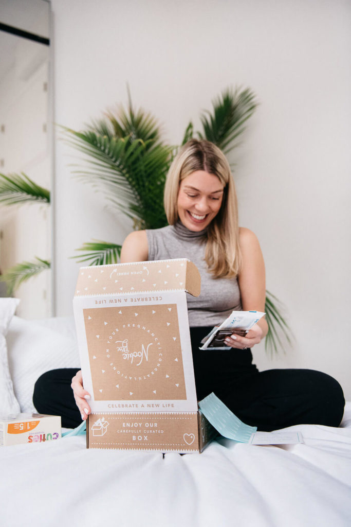 A woman smiling on a bed opens a subscription box, captured beautifully in brand photography. The brown box with white graphics is surrounded by small items, and a green plant adds a touch of nature in the background.