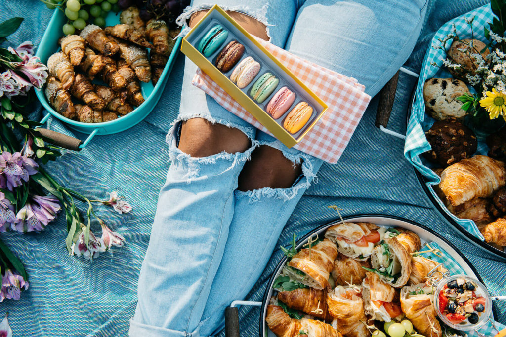 A person wearing ripped jeans sits on a blanket in a setting perfect for brand photography, surrounded by food like croissants, macarons, grapes, and flowers. The vibrant picnic spread includes a variety of snacks and a colorful bouquet, creating a cozy outdoor ambiance.