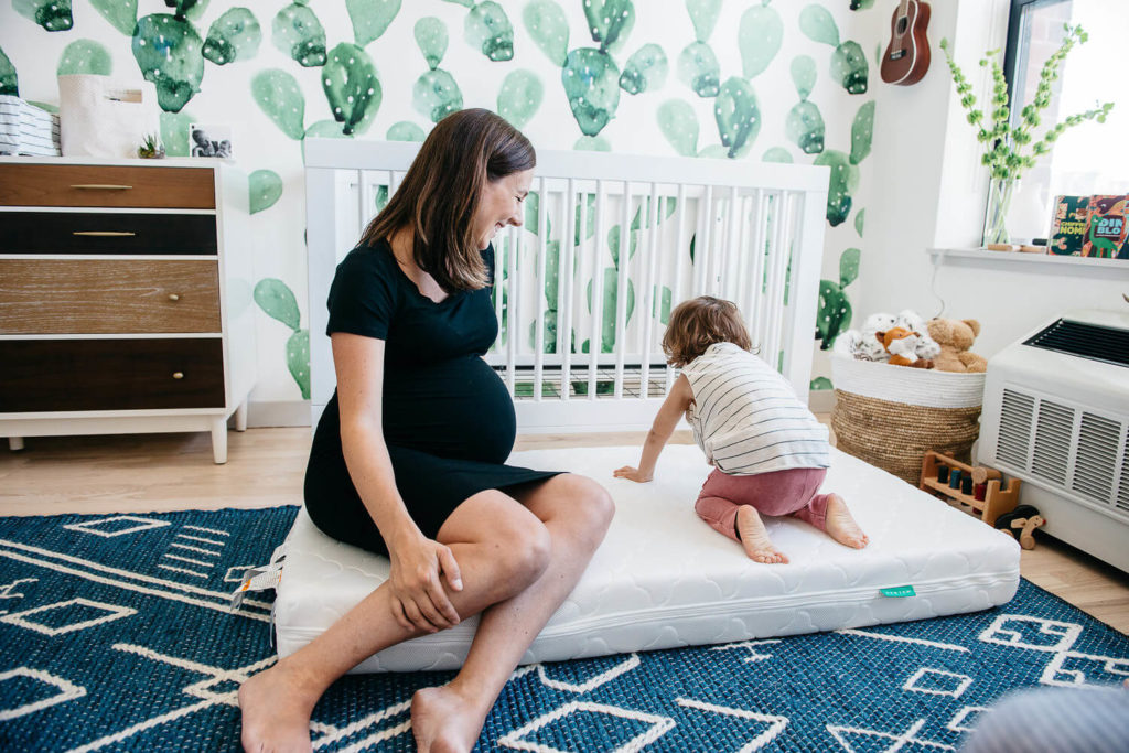 A pregnant woman sits on a crib mattress in the nursery, smiling at a young child playing nearby. The room, perfect for brand photography, features cactus-themed wallpaper, a dresser, and various toys.