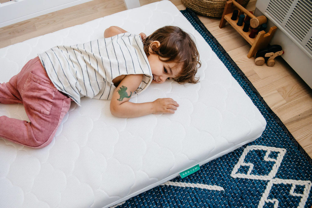 A toddler in a striped tank top and pink pants relaxes on a white mattress, embodying a moment perfect for commercial photography. Nearby, a toy and patterned blanket complete the cozy scene, while a temporary tattoo adds an adorable touch to this brand photography gem.