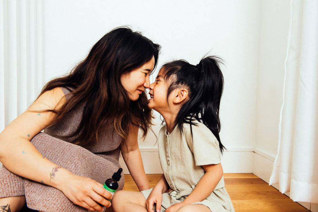 A woman and a child with long dark hair are sitting on a wooden floor, touching foreheads and smiling. In this moment of warmth and affection, the woman holds a small bottle with a dropper—a perfect example of brand photography capturing genuine connection.
