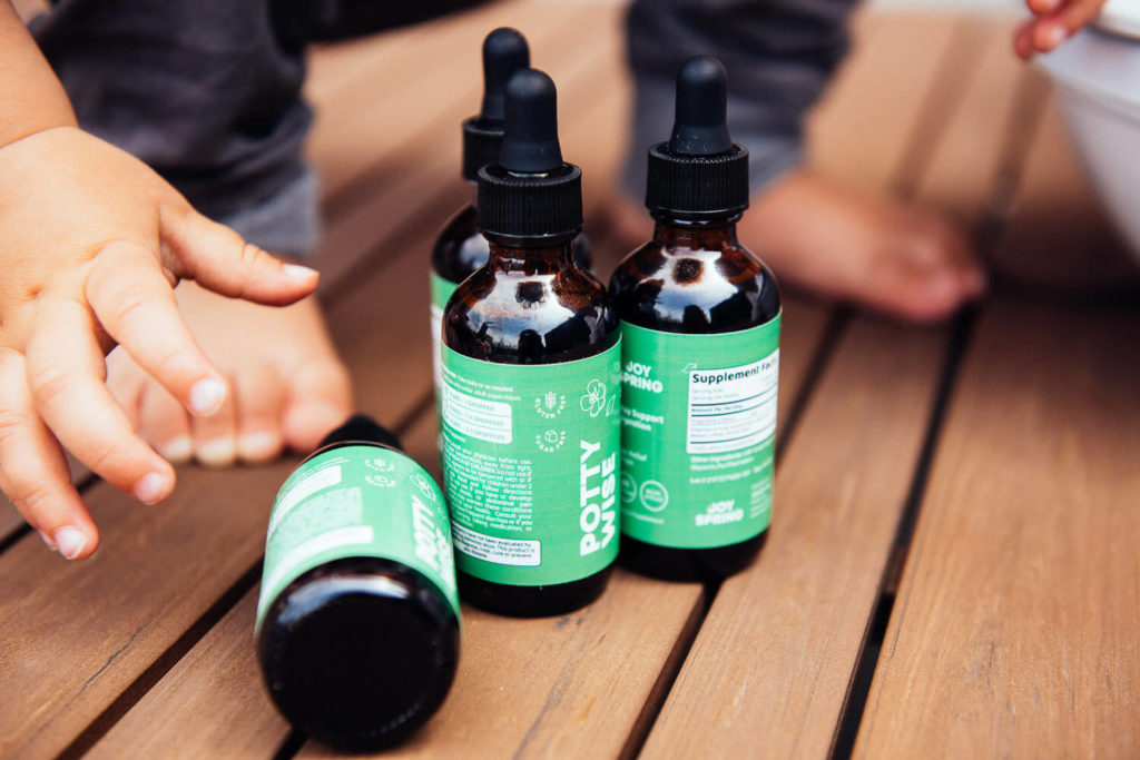 Four brown dropper bottles with green labels subtly capture the essence of brand photography as they rest on a wooden surface. A child's hand and feet reach towards the bottles, creating an inviting scene that blends warmth with the art of commercial photography.