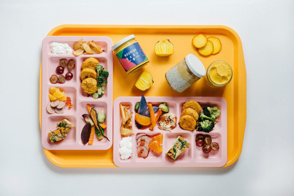 A vibrant cafeteria tray holds two pink meal trays brimming with assorted foods like veggies, fruit, and nuggets. Accompanied by two jars and a cupcake with yellow frosting, this stunning display of brand photography shines against the white background.