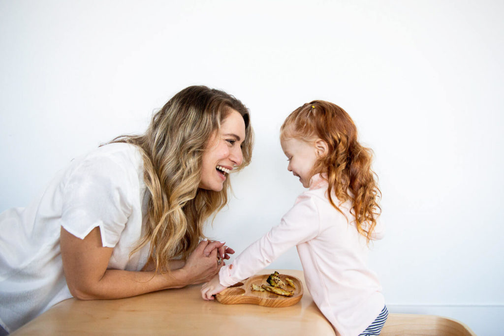 A woman with long hair laughs while interacting with a young girl with red hair. They lean over a wooden table with a plate of food, perfectly capturing the essence of brand photography against a plain white wall.