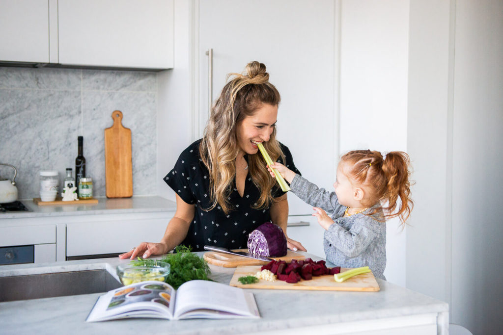 In a bright kitchen with white cabinets and marble elements, a woman and child engage in playful brand photography. The child stands on a stool, feeding the woman celery amid an array of vegetables and an open cookbook, capturing a moment perfect for commercial photography.
