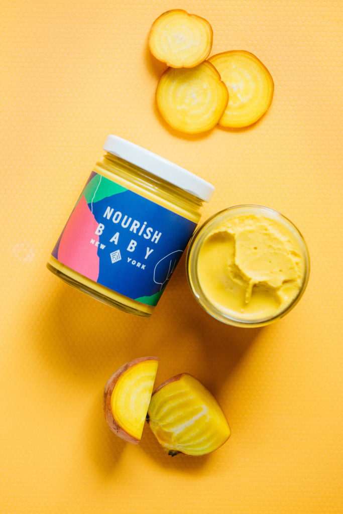 The brand photography showcases an open jar of Nourish Baby New York yellow baby food next to a halved golden beet. Set against a matching yellow surface, it creates a bright, monochromatic look that highlights the vibrant product.