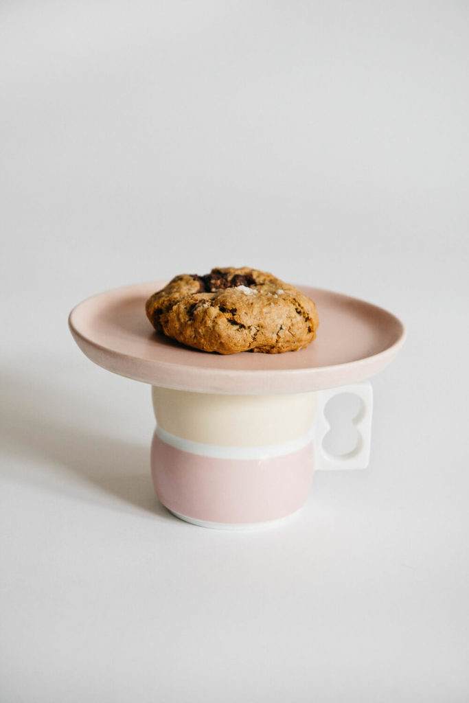 A single chocolate chip cookie rests on a light pink plate, balanced atop a two-toned pink and cream mug with a unique loop-shaped handle. This elegant setup is ideal for brand photography, set against a plain white background to highlight every detail.