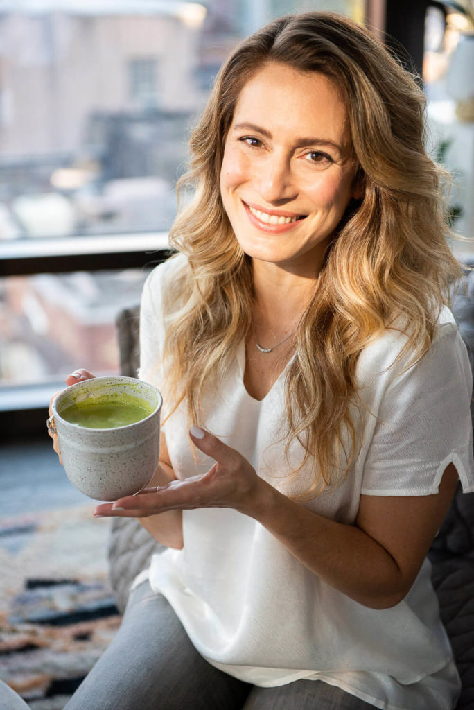 A person with long blonde hair, wearing a white shirt, is smiling and holding a cup of green tea. Framed in brand photography style, they are sitting by a window with an urban view in the background.