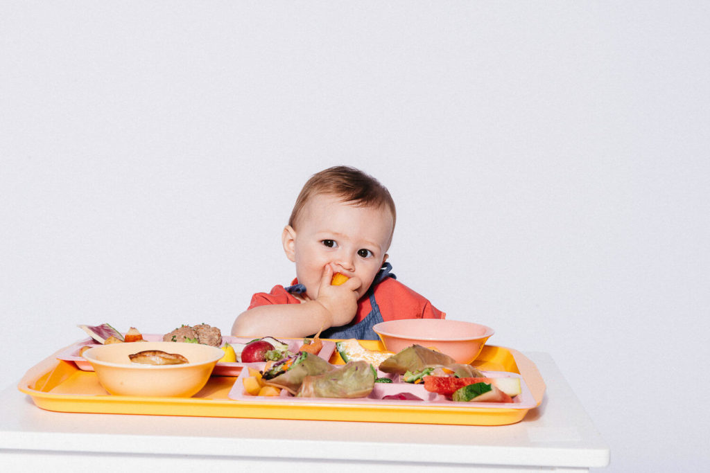 A baby sits at a table with a tray full of colorful food, captured in vibrant brand photography. The child is eating a piece of fruit with a thoughtful expression, surrounded by various small dishes of vegetables and snacks against a plain white background.
