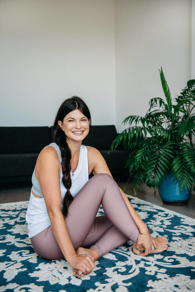 A woman with long dark hair, wearing a white tank top and pink leggings, sits cross-legged on a patterned blue and white rug. She is smiling indoors, ideal for brand photography, with a green plant and a dark sofa in the background.