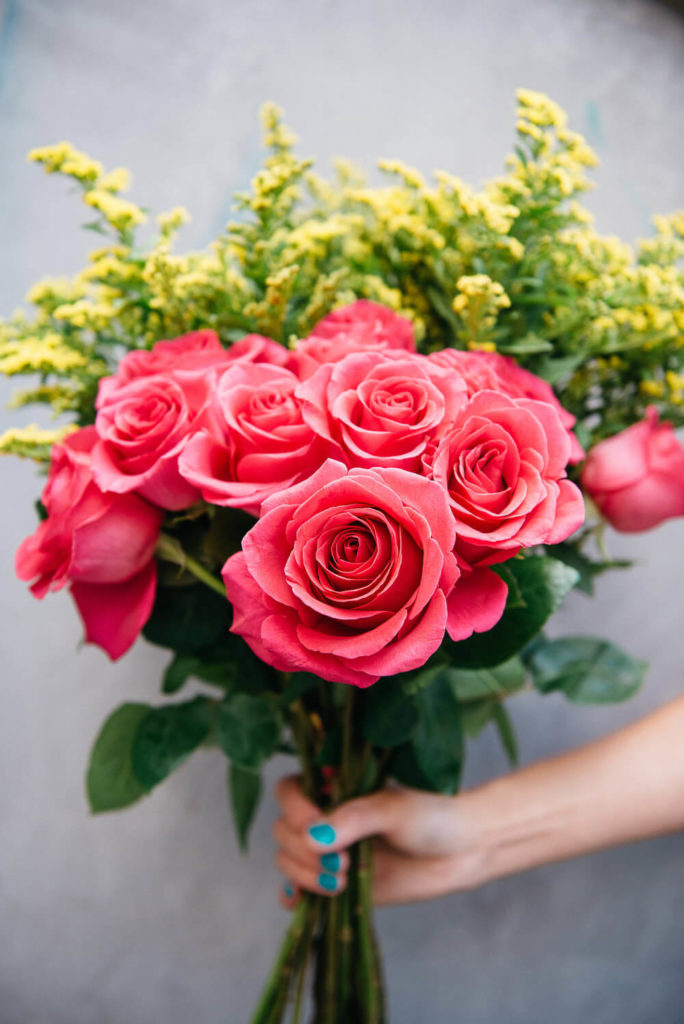 A bouquet of vibrant pink roses and yellow flowers is held against a plain background, perfect for commercial photography. A hand with painted blue-green nails grasps the stems, capturing brand photography elegance.