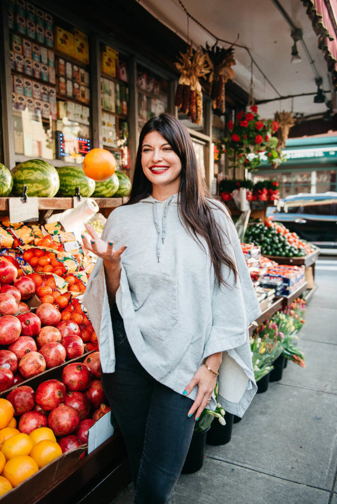 A woman in a gray poncho smiles amidst the vibrant stall, embodying the essence of brand photography. She holds leafy greens, surrounded by a commercial display of pomegranates, oranges, and watermelons. In the background, more market goods and flowers enhance the lively scene.