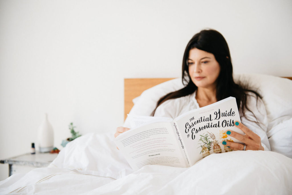 A person with long dark hair is sitting in bed, immersed in a book titled The Essential Guide to Essential Oils. The serene scene, perfect for brand photography, includes a bedside table adorned with a small vase, enhancing the calm and cozy setting.