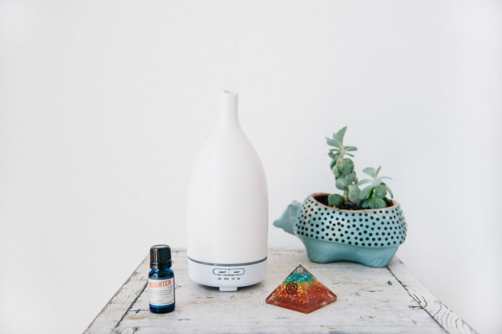 A minimalist scene ideal for brand photography features a white essential oil diffuser on a wooden table, accompanied by a small bottle of essential oil, a decorative pyramid, and a turtle-shaped planter with succulents against a plain white background.