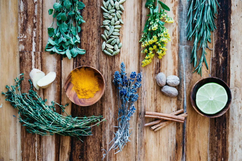 A rustic wooden table, perfect for brand photography, showcases an array of herbs and spices: fresh oregano, cardamom, rosemary, turmeric, nutmeg, cinnamon sticks, thyme, lavender, and a halved lime. Small bowls artfully hold some of the ingredients.