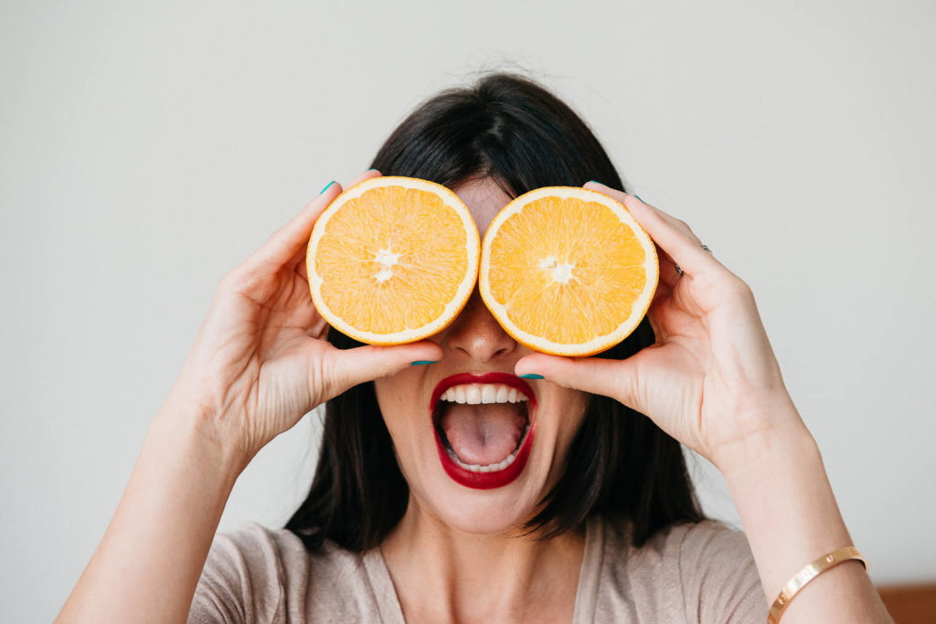 A woman playfully holds two orange halves over her eyes like glasses, with her mouth open wide as if laughing or shouting. Her nails are painted. The background is plain and light-colored, capturing a vibrant moment in brand photography.