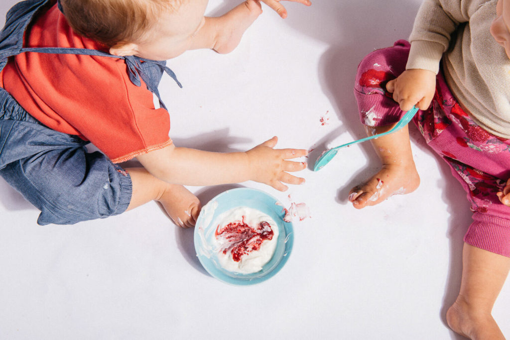 Two toddlers sit on the floor in a delightful display of brand photography, playing with a bowl of yogurt and red fruit sauce. One wears a red shirt and denim overalls, while the other sports a pink outfit. They happily make a mess with their hands and a blue spoon, capturing an authentic moment.