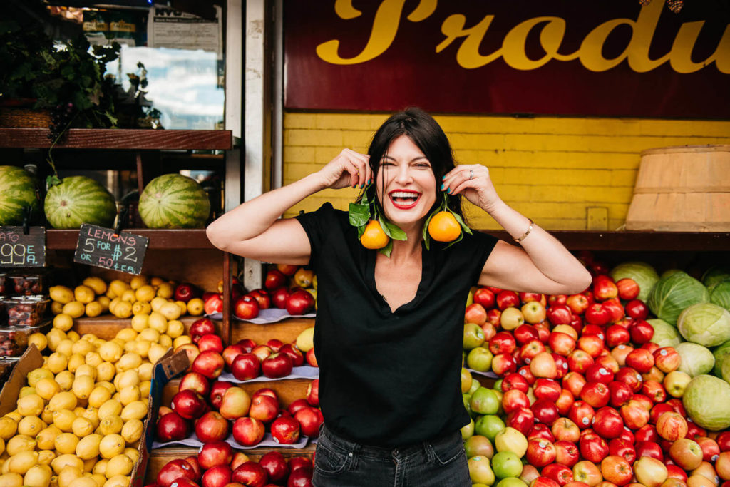 A person playfully holds two pears up to their ears in a lively display of brand photography at an outdoor market. Surrounded by apples, limes, and watermelons, they smile broadly in their black shirt, perfectly capturing the vibrant essence of commercial photography.