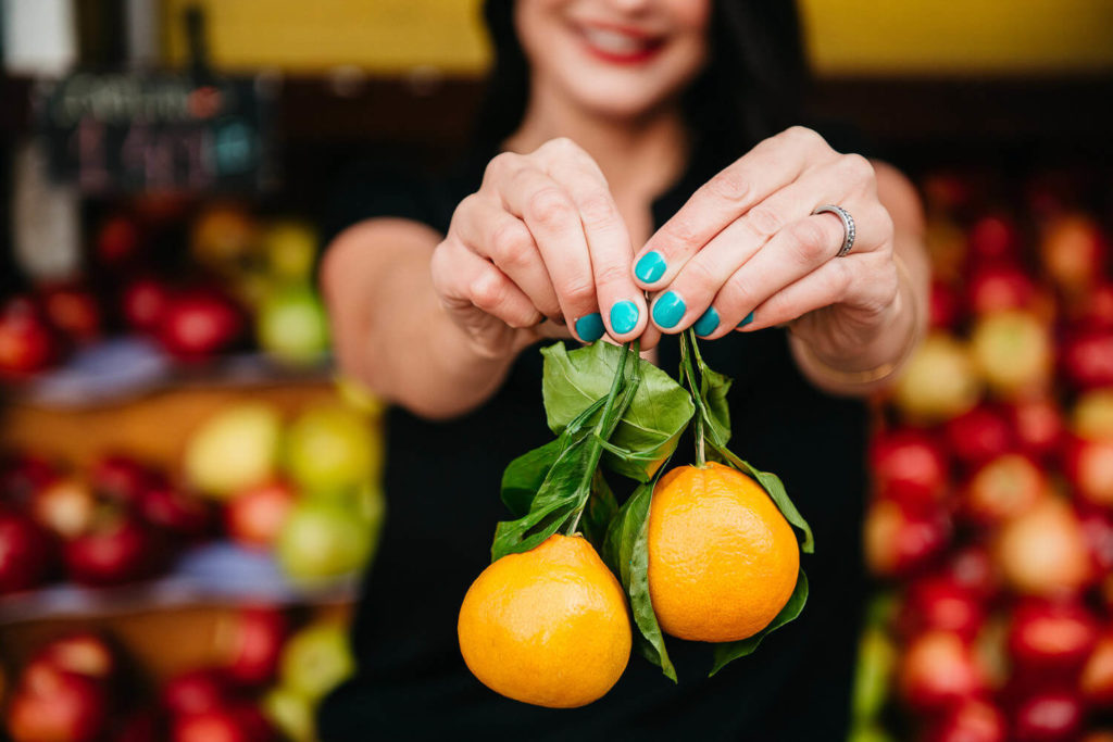 A woman with blue-painted nails holds two oranges with green leaves, exuding a vibrant energy perfect for brand photography. She smiles against a backdrop of diverse fruits, including apples, while the focus remains on the oranges in this captivating scene.