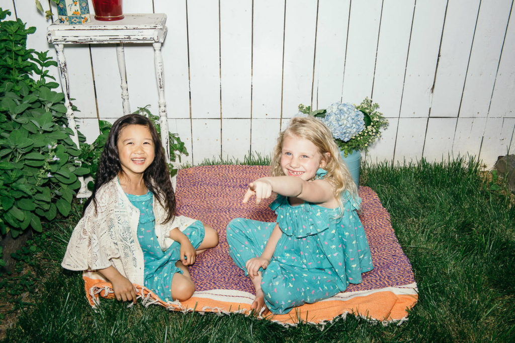 Two young girls sit on a colorful blanket in a grassy backyard, perfect for brand photography. Both are smiling and wearing matching turquoise dresses. One is pointing forward. Behind them, a small white table holds a red and blue bouquet, with a white wooden fence forming the backdrop.