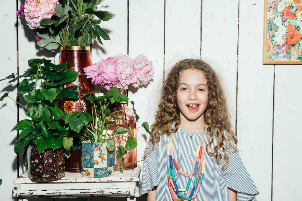 A smiling child with curly hair stands in front of a white wooden wall, embodying the essence of brand photography. They wear a striped shirt and colorful necklace, surrounded by vibrant pink flowers and green plants in decorative pots.