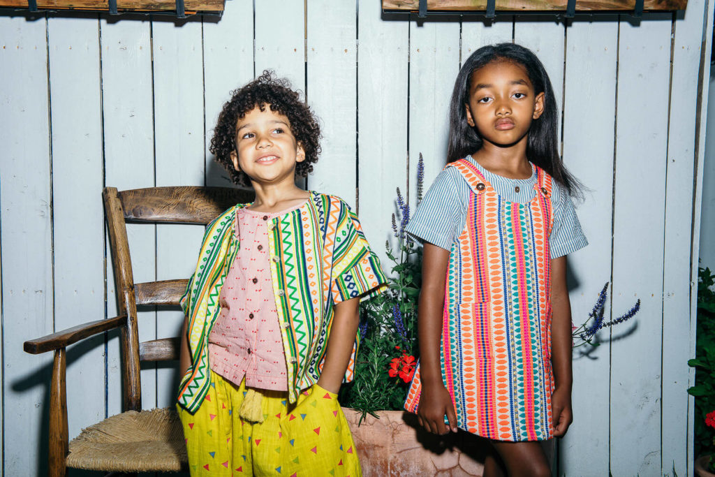 Two children pose in front of a white wooden wall, embodying the joy and vibrancy captured in brand photography. The child on the left sports curly hair, a colorful striped jacket, and yellow pants, while the one on the right wears a patterned dress. Nearby, a wooden chair and flowers complete this commercial photography scene.