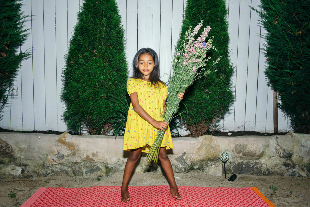 A young girl in a yellow dress stands on a red patterned mat, holding a large bouquet of pink and white flowers. She is barefoot with a serious expression, embodying the charm often captured in brand photography. Three tall, lush trees rise behind her against a wooden fence.