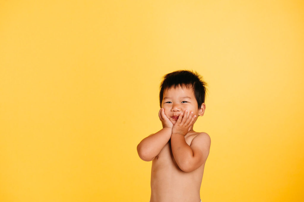 A young child with short black hair stands against a bright yellow background, embodying the essence of brand photography. The child is smiling with hands pressed to their cheeks, expressing surprise or joy, capturing the magic often sought in commercial photography.