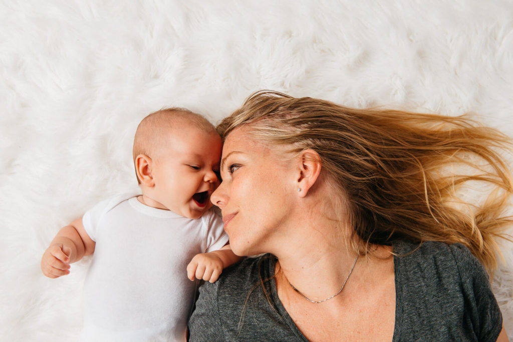 A baby in a white onesie lies on a fluffy white surface, next to a woman with long hair. Both are facing each other, with the woman smiling gently and the baby appearing to talk or laugh—a perfect moment captured in brand photography.