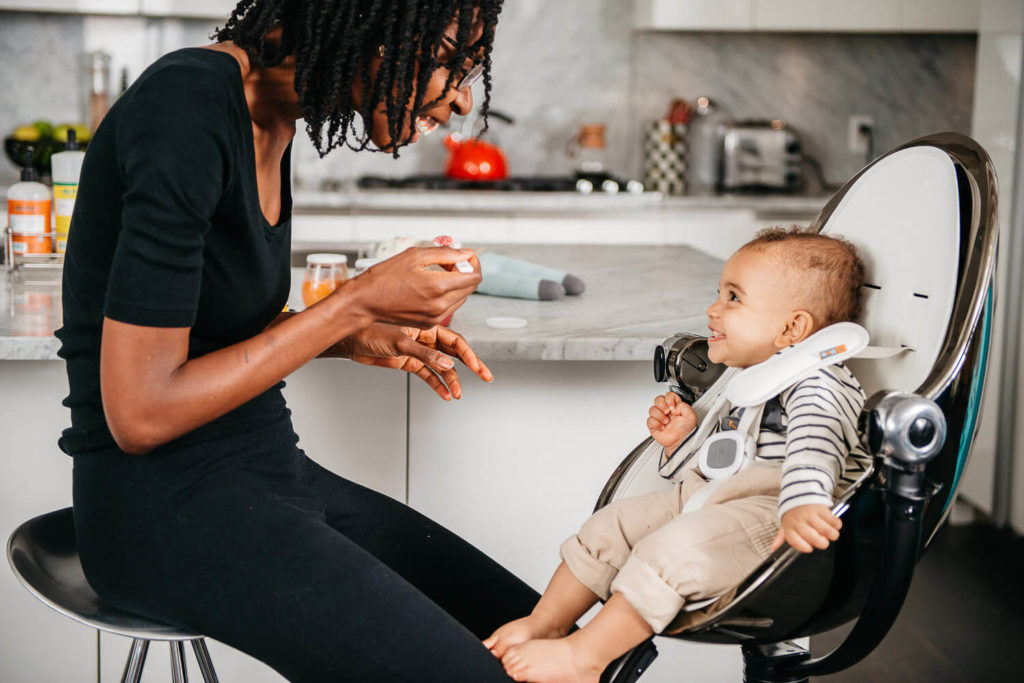 A woman is feeding a smiling baby sitting in a high chair in a modern kitchen, showcasing the essence of brand photography. She's holding a spoon and a small food container, with various kitchen items adorning the countertop in the background.