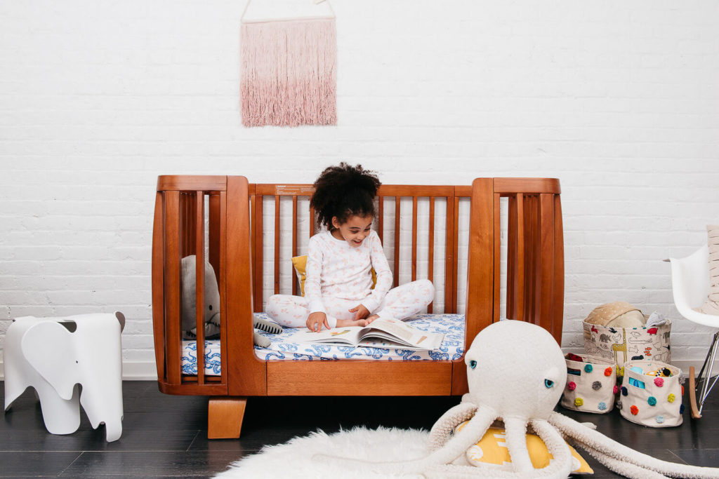 A child is sitting on a wooden toddler bed, absorbed in a book. The room, styled for brand photography, features a plush octopus, an elephant-shaped chair, and baskets. A white rug graces the floor while a pink wall hanging adds charm against the white brick backdrop.