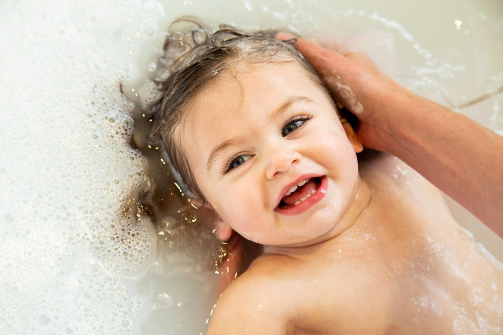 A smiling baby enjoys a bath, surrounded by soapy water, as an adult's hands gently wash their hair. The scene captures the essence of commercial photography, with the baby looking happy and relaxed amid the shimmering bubbles.