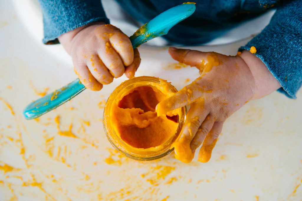 A child’s hands, covered in orange puree, grasp a small jar and a blue spoon, embodying the essence of brand photography. The puree is playfully spread across the white surface, while the child wears a blue long-sleeve shirt—a perfect capture for commercial photography.