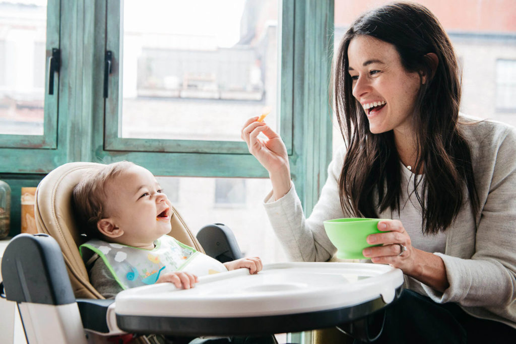 In a moment perfect for commercial photography, a woman with long dark hair smiles as she feeds a laughing baby in a highchair. The baby, wearing a bib, giggles while the woman holds a green bowl. They sit by large windows offering views of surrounding buildings, capturing pure happiness.