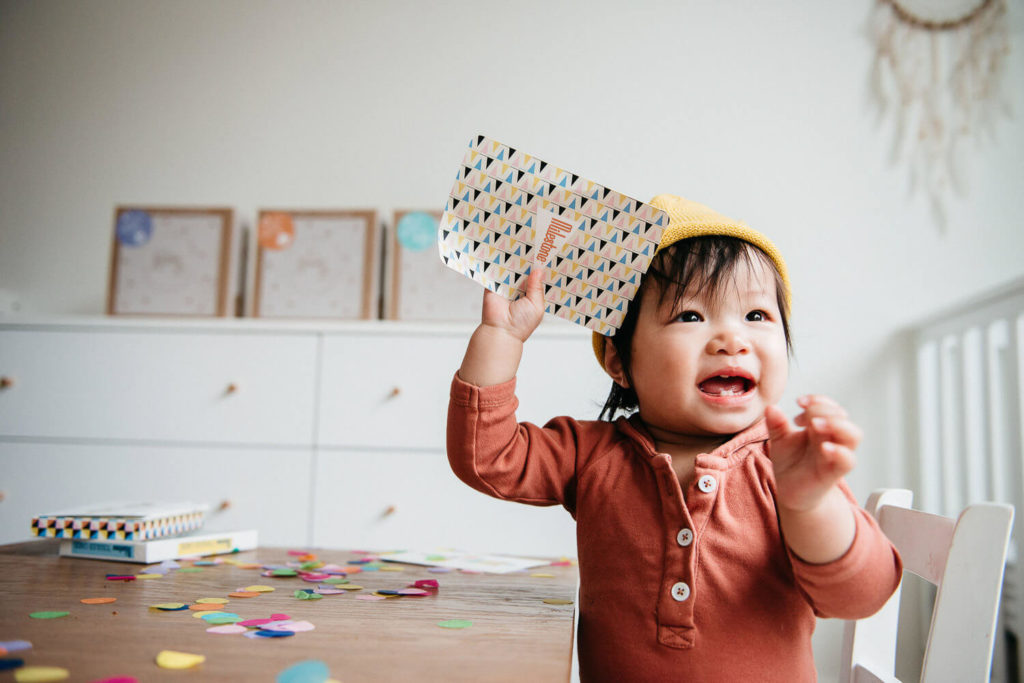 A smiling toddler in a yellow hat and red shirt holds a colorful book at a confetti-covered table, embodying the vibrancy of commercial photography. The room's white dresser and festive decorations complete the brand photography scene, creating an inviting atmosphere.