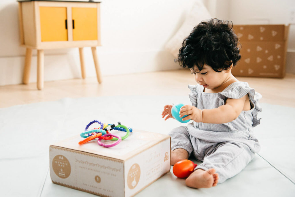 A toddler with curly hair sits on a light blue mat, playing with colorful toys. The child clutches a blue ball next to a cardboard box, framed perfectly for brand photography. A wooden cabinet with a yellow door is visible in the background, adding charm to the scene.
