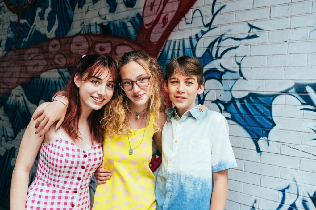 Three young people stand close together, smiling in front of a colorful mural featuring a large octopus and waves. Captured by a Brooklyn family photographer, the individual on the left wears a pink checkered dress, the middle sports yellow attire, while the right is in a blue and white shirt.