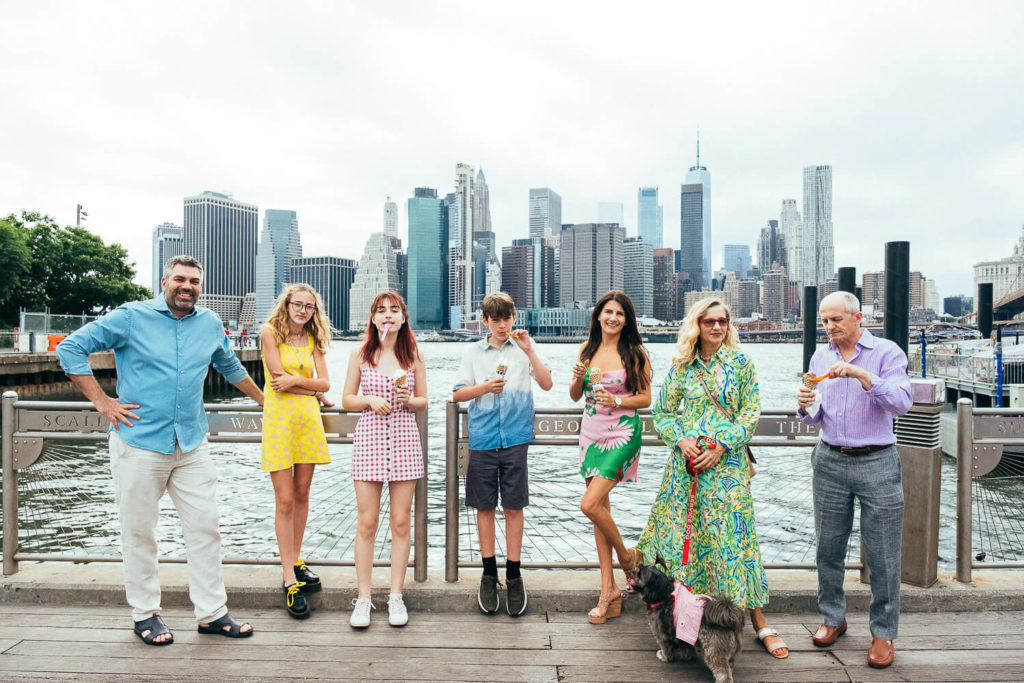 A group, possible subjects for a family photography NYC session, including three adults and three teenagers, stand on a pier with ice cream cones. The city skyline and waterfront are visible in the background. One person has a dog on a leash. They are dressed in summer clothing.