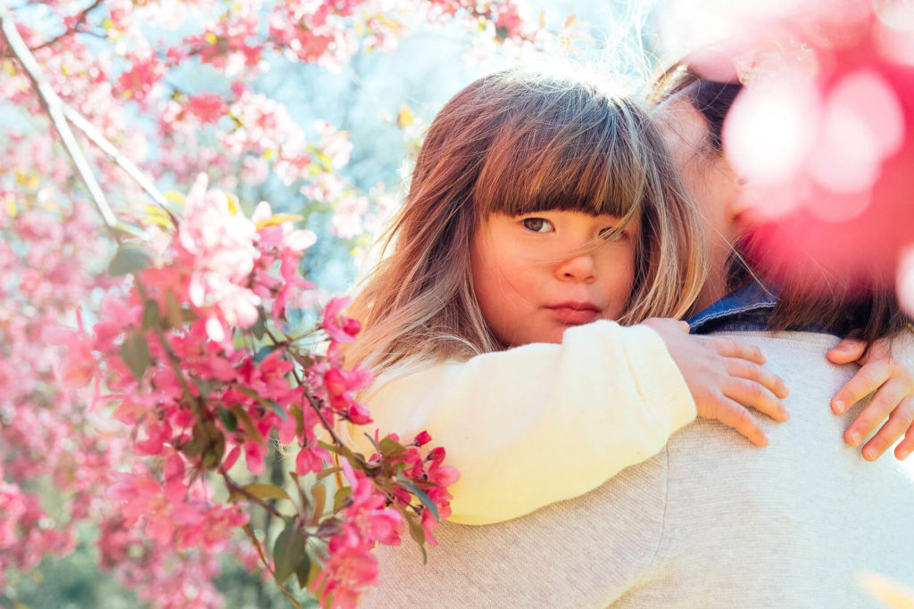 A child with long hair and a yellow sweater is held by an adult amidst vibrant pink blossoms on a sunny day. Captured by a Brooklyn family photographer, the serene expression as they look into the camera adds to the charm of this family photography NYC moment.