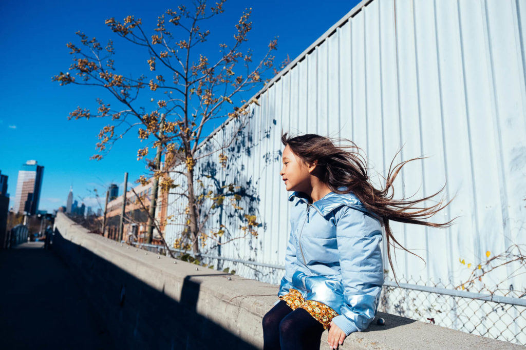 young girl in a light blue jacket sitting on a ledge with a backdrop of autumn foliage and a bright sky