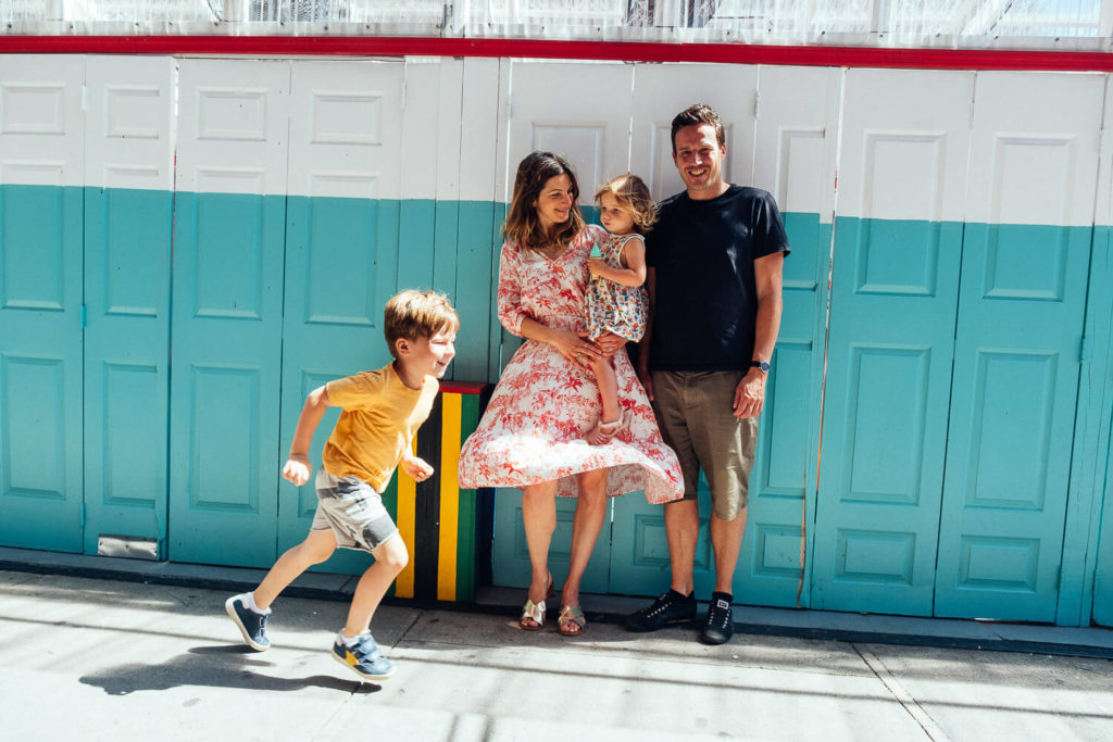 A family stands in front of a colorful wall with a blue and white pattern. A woman holds a young girl, while a man stands beside them. Captured by a family photography NYC expert, a young boy in a yellow shirt joyfully runs past them on the sidewalk.