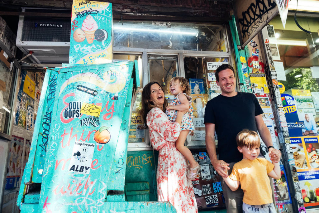 A cheerful family stands outside a colorful, graffiti-covered shop. Captured by a talented Brooklyn family photographer, the scene shows the mother in a floral dress holding a child, while the father in a black shirt holds hands with another. Bright posters and stickers complete this vibrant NYC family moment.