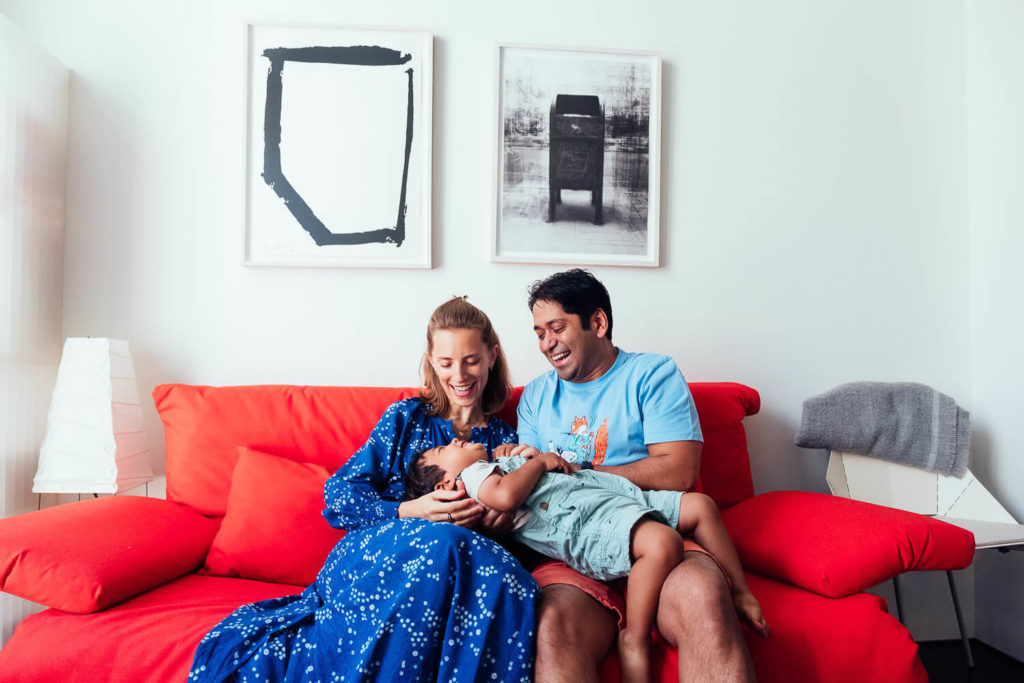 A family of three sits on a bright red couch, captured beautifully by a Brooklyn family photographer. The mother, in a blue dress, and the father, in a light blue shirt, joyfully interact with their young child lying across their laps. Two abstract art pieces hang on the wall behind them.