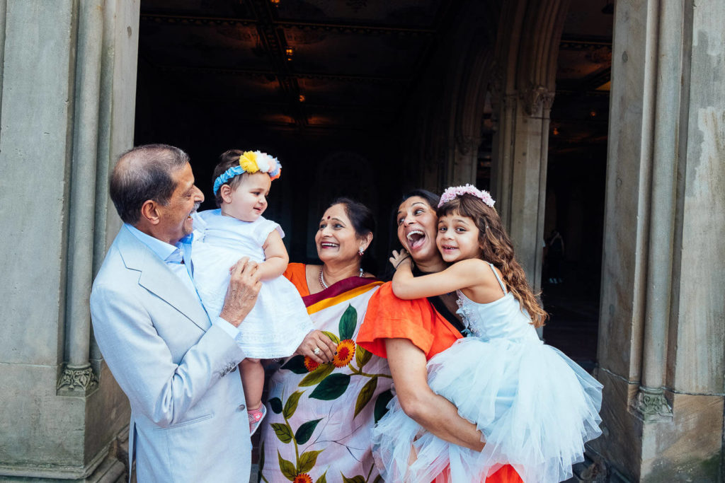 A joyful family moment captured by a Brooklyn family photographer shows two adults holding young girls in white dresses and floral headbands. They stand in an arch, sharing smiles and laughter. The adults, clad in formal attire with one wearing a sari, create a timeless scene of joy amidst an ornate setting.