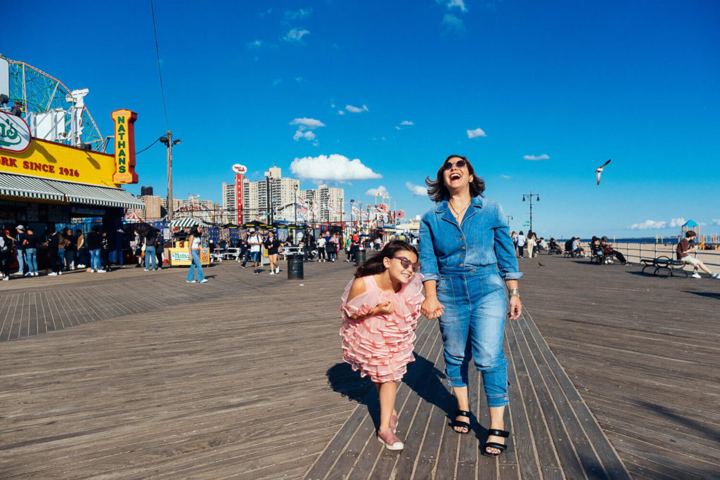 A woman and a girl, captured by a talented family photographer in NYC, hold hands and laugh while strolling on a sunny boardwalk. The sky is blue with a few clouds, and people gather near shops and attractions in the background.